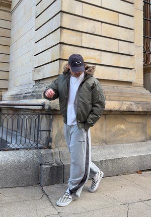 Cazadora bomber verde con capucha forrada de piel, camiseta blanca, pantalones de chándal grises con rayas negras laterales y zapatos de cuadros en blanco y negro.