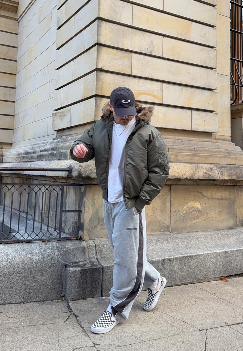 Grüne Bomberjacke mit pelzgefütterter Kapuze, weißes T-Shirt, graue Jogginghose mit schwarzen Seitenstreifen und schwarz-weiß karierte Schuhe.