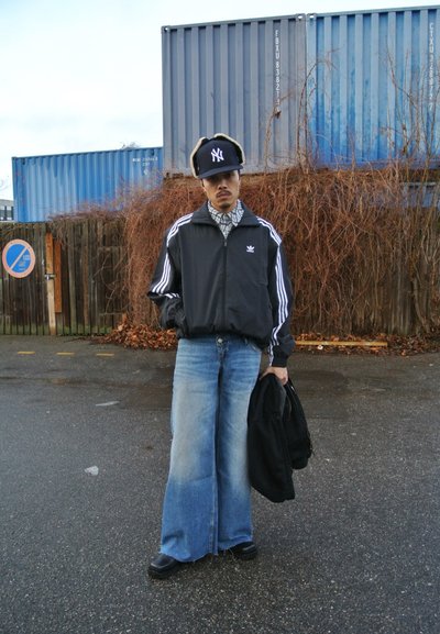 Chaqueta negra con cremallera y líneas blancas, camisa de cuadros, jeans de pierna ancha, zapatos negros y una gorra negra con un logo, de pie frente a una valla.