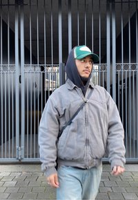 Giacca bomber grigia con cappuccio, jeans blu e berretto verde. Camminando vicino a un cancello metallico in un'area pavimentata texturizzata.