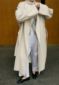 Personne portant un long manteau beige, un pantalon de survêtement gris clair, un cardigan gris et des chaussures noires à enfiler, debout sur un sol gris devant un mur en bois.