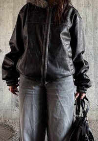 Osoba ve černé kožené bundě s kožešinovým límcem, šedých kalhotách, držící černou kabelku před pozadím drsné betonové zdi.