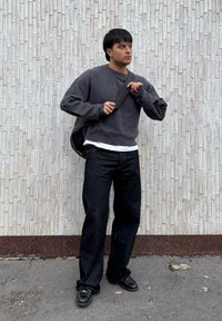 Νεαρός άνδρας που προσαρμόζει το λουρί μαύρης τσάντας ώμου, φοράει σκούρο γκρι πουλόβερ, μαύρο παντελόνι και μαύρα παπούτσια, στέκεται μπροστά σε υφή λευκού τοίχου.