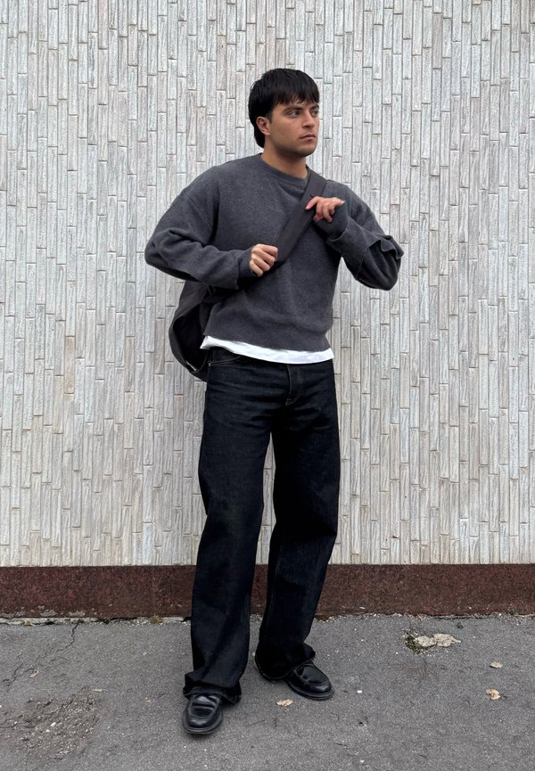 Mladić podešava remen crne torbe preko ramena, nosi tamnosivi džemper, crne hlače i crne cipele, stojeći uz teksturirani bijeli zid.
