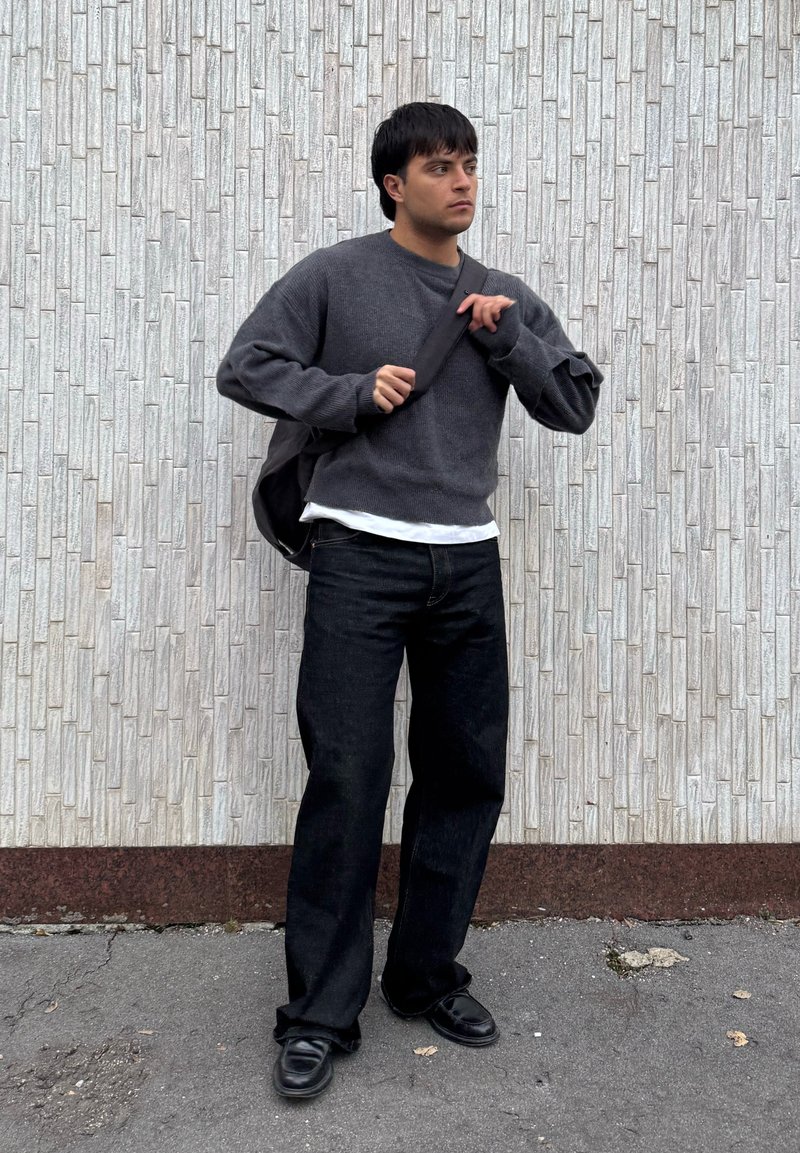 Jeune homme ajustant la sangle de son sac à bandoulière noir, portant un pull gris foncé, un pantalon noir et des chaussures noires, debout devant un mur blanc texturé.