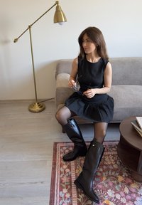 Kvinna i svart dress med ny stripes och höga svarta stövlar sitter på en grå soffa och håller i solglasögon, bredvid en mässingsgolvlampa och en blommig matta.