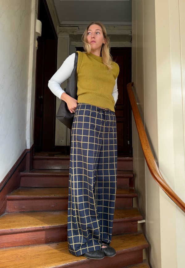 Mujer de pie en unas escaleras de madera, vistiendo un chaleco mostaza, camisa blanca de manga larga, pantalones anchos con cuadros, zapatos negros y llevando un bolso negro.