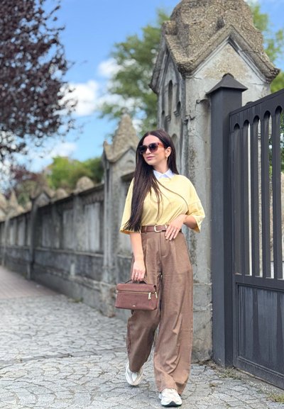 Top amarillo de gran tamaño, pantalones anchos marrones y zapatillas blancas; sosteniendo un pequeño bolso marrón. Pelo largo y liso y gafas de sol negras. Entorno al aire libre.
