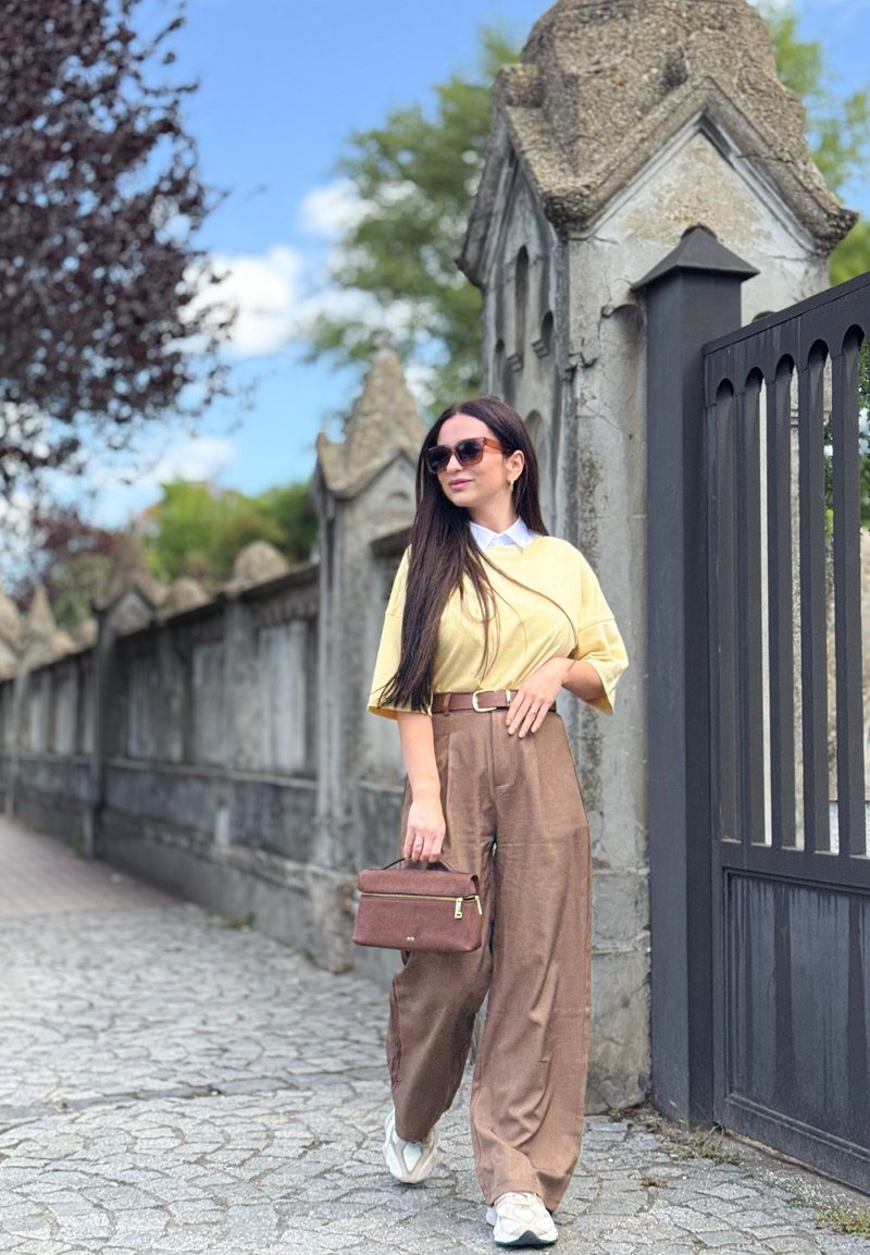Camisola oversized amarela, calças largas castanhas e sapatilhas brancas; segurando uma pequena mala castanha. Cabelo liso e comprido e óculos de sol pretos. Ambiente ao ar livre.