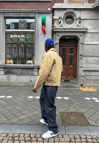 Beige bomberjack, losse donkere jeans, witte sneakers met een blauwe Nike swoosh. Stedelijke straatsetting bij een winkel met een stenen façade.