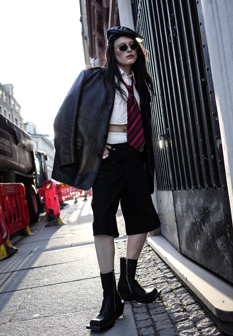 Personne appuyée contre une grille métallique noire porte une veste en cuir noire sur les épaules, une chemise blanche courte, une cravate rayée, un short noir, des bottes et des lunettes de soleil.