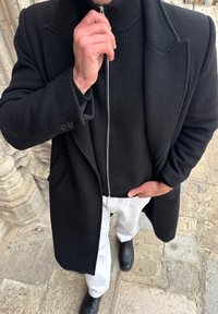 Personne en manteau noir, pull à fermeture éclair noir, pantalon blanc et chaussures noires, debout sur un pavé en pierre, une main dans la poche, l'autre main près du visage.