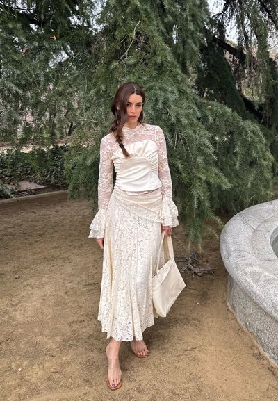 Mujer con un vestido largo de encaje blanco y un bolso crema, de pie descalza en un camino de tierra cerca de un árbol perenne y un banco de piedra.