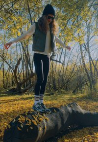 Žmogus stovintis ant rąsto, dėvintis žalią liemenę, baltą ilgomis rankovėmis marškinį, juodus leginsus ir žalius sportbačius. Aplink rudens lapai.