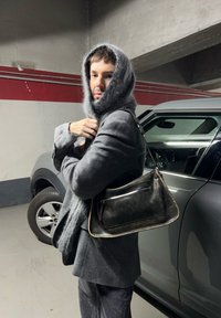 Bolsa de couro preta com alça para o ombro; xale cinza suave usado com um blazer cinza escuro. Fundo de uma garagem de estacionamento em betão.