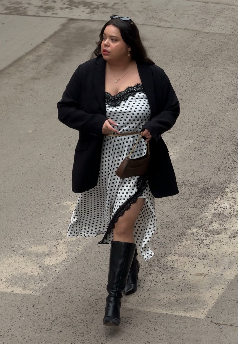 Femme marchant dans la rue portant un blazer noir, une robe blanche à pois noirs avec dentelle noire, des bottes noires, tenant un sac à main marron, regardant vers sa gauche.