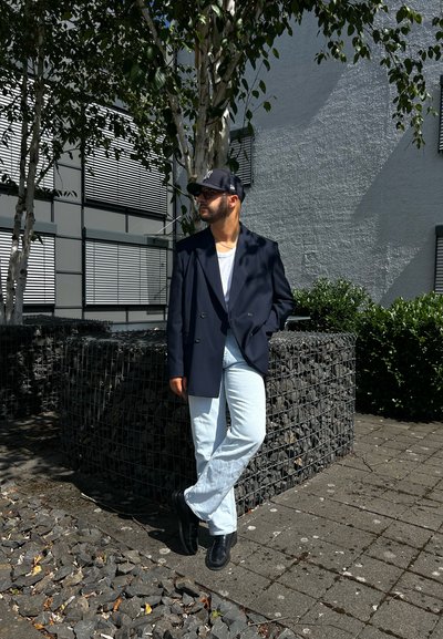 Blazer azul marino de doble botonadura, camiseta blanca, jeans de color azul claro y zapatos negros. Situado frente a un edificio moderno con vegetación y acentos de piedra.