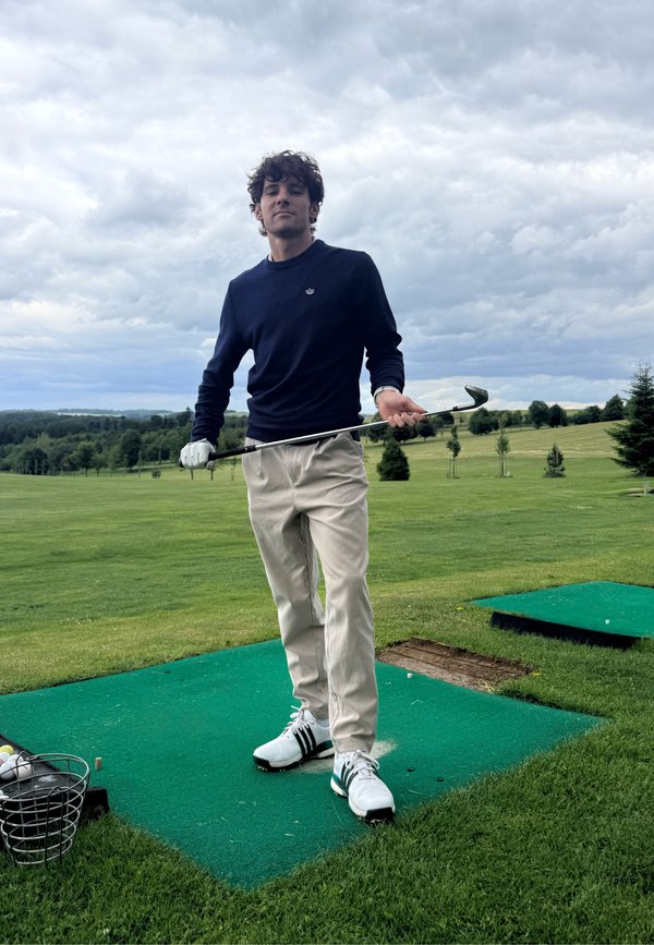 Nuori mies golfhansikkaassa pitää golfmailaa harjoitusviheriöllä, jossa kori täynnä golfpalloja, pilvinen taivas ja taustalla puita.