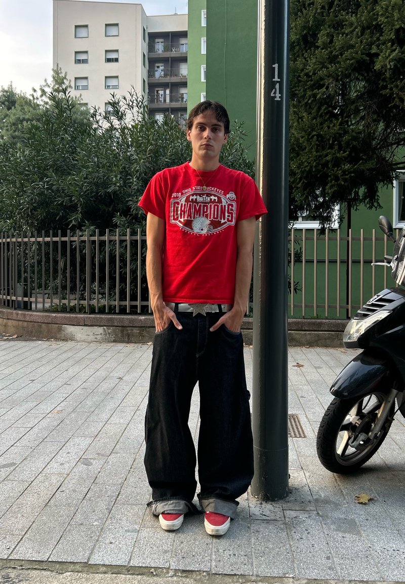 Red t-shirt with "Champions" graphic, black baggy jeans, and red sneakers. Standalone against a backdrop of plants and buildings.