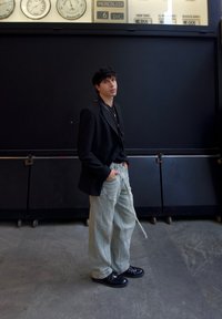 Blazer nero a doppio petto sopra una camicia nera, pantaloni larghi verde chiaro e scarpe nere. Lo sfondo presenta orologi e un muro scuro.