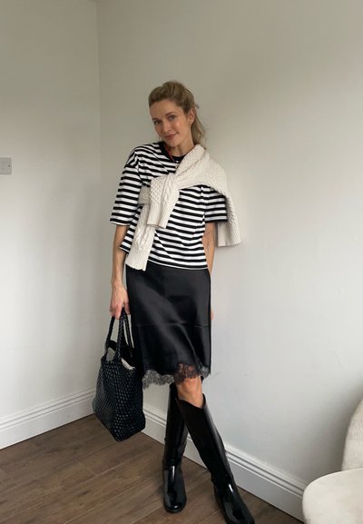 Mujer con blusa de rayas negras y blancas, falda negra con borde de encaje, botas negras y un suéter blanco sobre los hombros, sosteniendo un bolso negro.