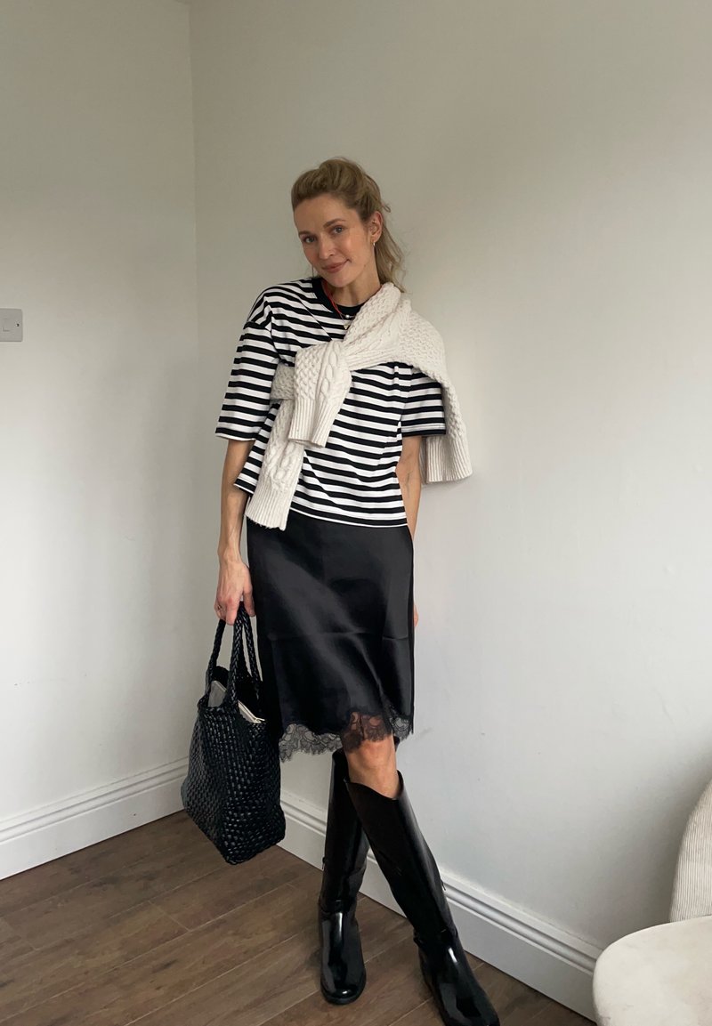 Femme portant un haut rayé noir et blanc, une jupe noire avec un bord en dentelle, des bottes noires, et un pull blanc drapé sur ses épaules, tenant un sac noir.