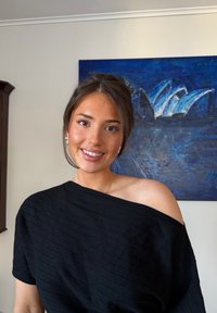 Jeune femme aux cheveux bruns attachés, portant un haut noir à épaules dénudées, souriant devant une peinture abstraite bleue sur un mur beige.