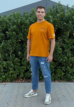 Camiseta de algodón naranja con estampado gráfico; vaqueros claros deshilachados; zapatillas blancas con detalles en beige; de pie al aire libre.