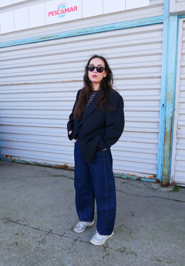Γυναίκα με γυαλιά ηλίου, σκούρο σακάκι, ριγέ πουκάμισο, φαρδιά τζιν και λευκά αθλητικά παπούτσια, στέκεται με τα χέρια στις τσέπες δίπλα σε μεταλλική περσίδα με την επιγραφή «Pescamar».
