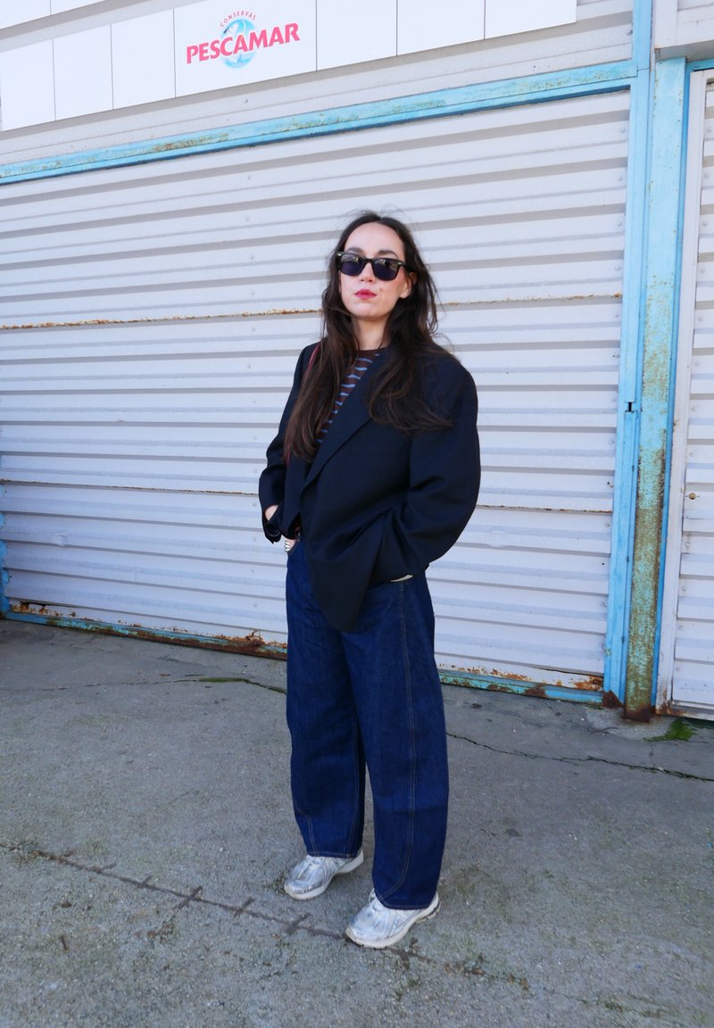 Femeie purtând ochelari de soare, sacou închis la culoare, cămașă în dungi, blugi largi și adidași albi, stând cu mâinile în buzunare lângă o jaluzea metalică cu semnul „Pescamar”.