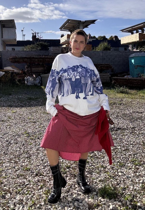 Une personne pose sur un chantier, vêtue d’un sweat-shirt blanc à motif graphique, d’une jupe rouge à carreaux et de bottes noires.