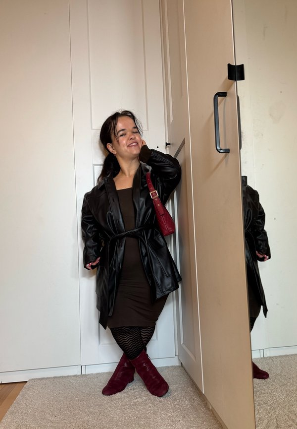 Schwarze Lederjacke mit Gürtel über einem braunen Kleid, kombiniert mit burgunderfarbenen Stiefeln. Schwarze gemusterte Strumpfhosen sichtbar. Steht neben einem Spiegel.