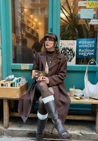 Brun oversized jakke, grå genser, rutete skjørt, gjennomsiktige strømpebukser, brun høye støvler med spenne. Stabler av bøker, kaffe og et bord ute.