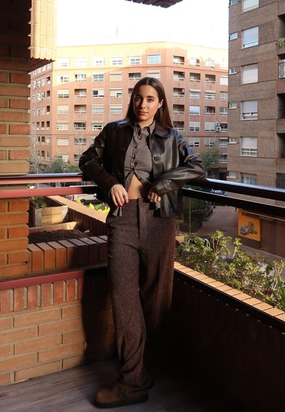 Chaqueta de cuero negra, camisa de rayas, pantalones marrones texturizados, de pie en un balcón con barandilla de ladrillo y plantas en cajas de madera.