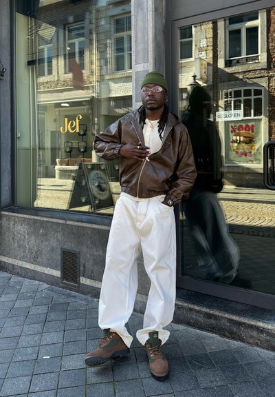 Hombre con gorro verde, chaqueta de cuero marrón, pantalones blancos y botas, de pie en la acera de una ciudad frente a una tienda con escaparate de cristal y reflejos.