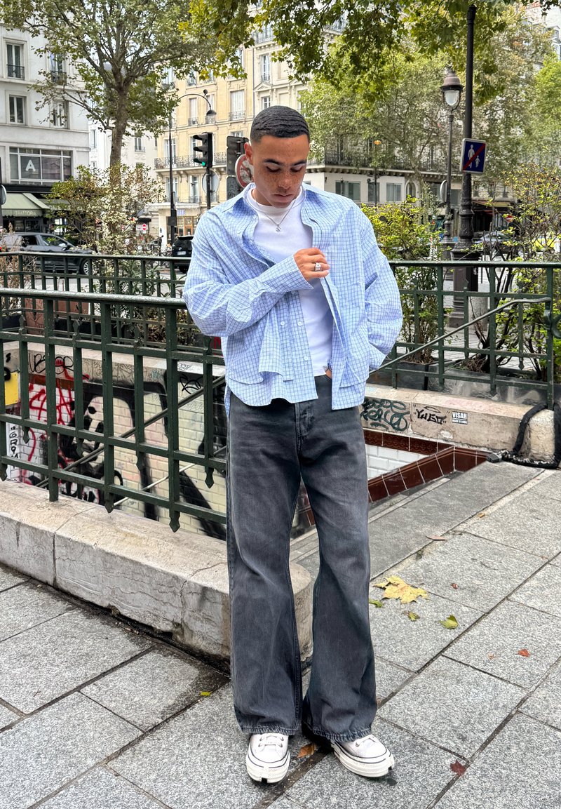 Camicia a quadri azzurro chiaro sopra una maglietta bianca, abbinata a jeans grigi scuri larghi e sneakers bianche. Sfondo urbano con vegetazione.