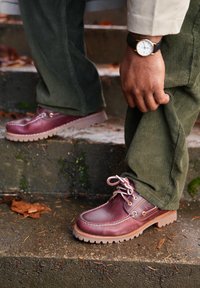 Brown leather boat shoes with white laces and rubber sole. Visible stitching detail; paired with green corduroy trousers on outdoor steps.