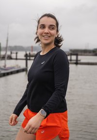 Μαύρο αθλητικό μπλουζάκι με μακριά μανίκια και στρογγυλή λαιμόκοψη συνδυασμένο με φωτεινές πορτοκαλί βερμούδες τρεξίματος. Φοράει ρολόι στον αριστερό καρπό.