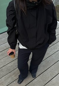 Jachetă ușoară neagră cu guler înalt, asociată cu o cămașă albă și pantaloni negri. Ținând o cană de cafea maro. Fundal din suprafață de lemn.