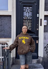 Felpa marrone con "WYOMING" in giallo, con un grafiche di cowboy. Shorts di jeans e una porta scura con decorazioni in metallo nero nel background.