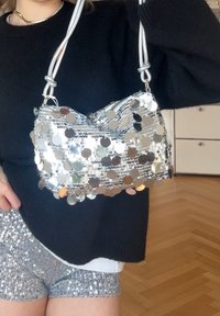 Sac à paillettes argenté en forme de pochette, avec une finition métallique et des bretelles en corde tressée. Le sac est orné de disques ronds réfléchissants.