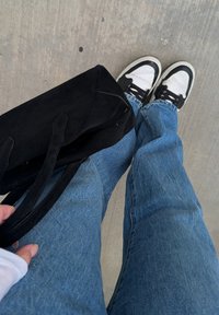 Personne portant un jean bleu et des baskets noir et blanc tenant un sac fourre-tout noir sur un sol en béton gris.