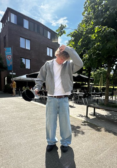 Sudadera gris con cremallera sobre una camiseta blanca, vaqueros de mezclilla claros y sueltos, zapatos negros, de pie al aire libre con árboles y un edificio al fondo.