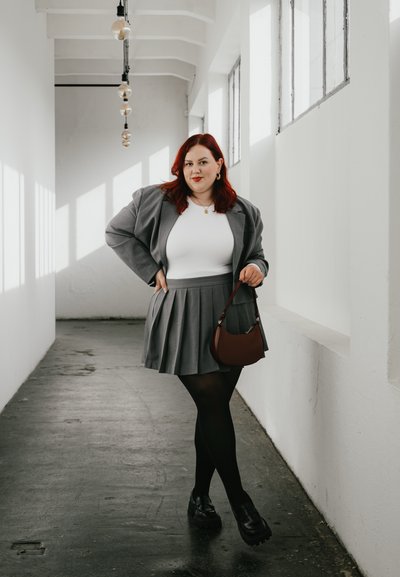Blazer gris sobre una blusa blanca, combinado con una falda gris plisada. Medias negras y zapatos negros gruesos. Un bolso marrón realza el conjunto.