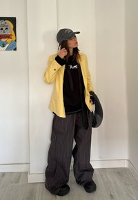 Personne en veste jaune, chemise noire, pantalon large rayé, casquette grise, tenant un chapeau gris, debout contre un mur blanc avec une petite œuvre d'art colorée.