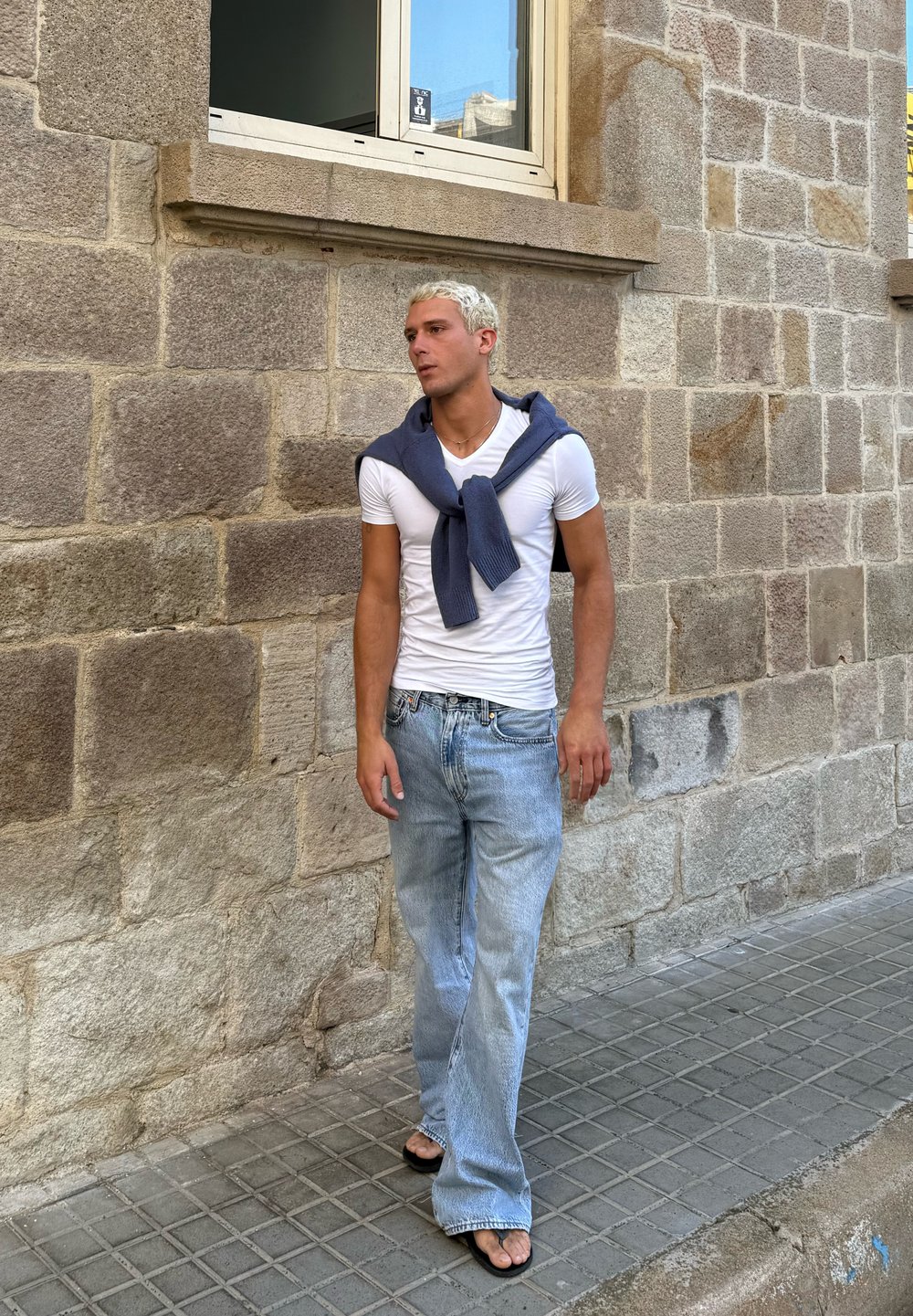 Bela prilagojena majica, svetlo modri flare jeans, in črne japonke. Temno modra jopica je ohlapno povezana okoli ramen. Ozadje iz kamnite stene.