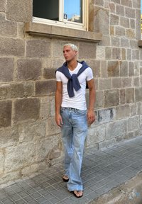 Bela prilagojena majica, svetlo modri flare jeans, in črne japonke. Temno modra jopica je ohlapno povezana okoli ramen. Ozadje iz kamnite stene.