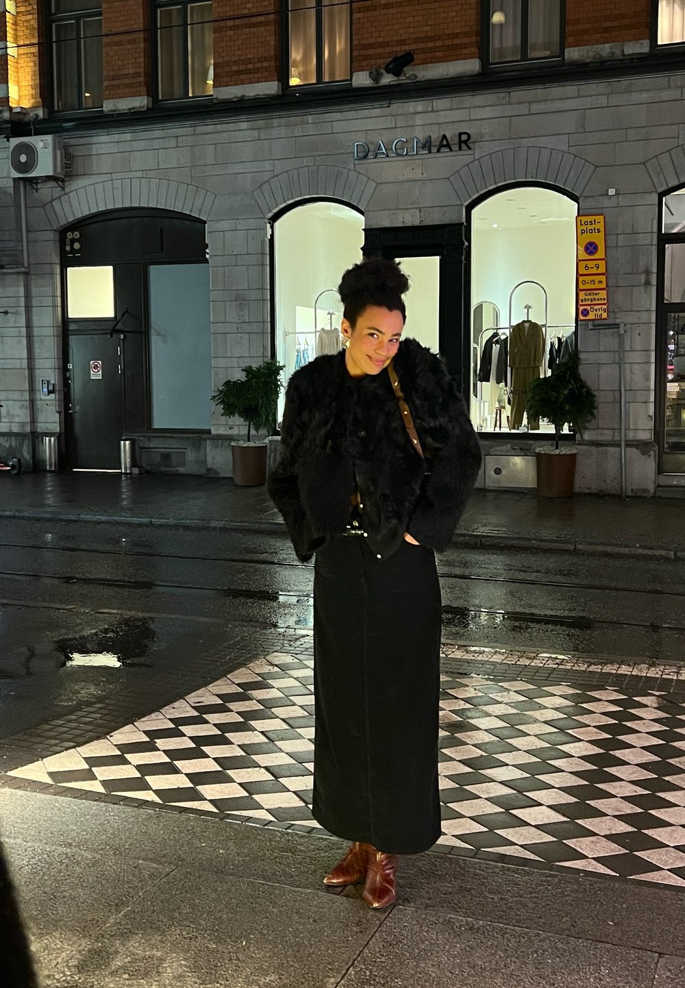 Μαύρο, φουσκωτό μπουφάν από ψεύτικη γούνα πάνω από μια μαύρη φούστα. Καφέ μποτάκ�ια μέχρι τον αστράγαλο και τσάντα με χρυσό λουράκι. Πλακόστρωτο σε μορφή καρό, αστικό φόντο.