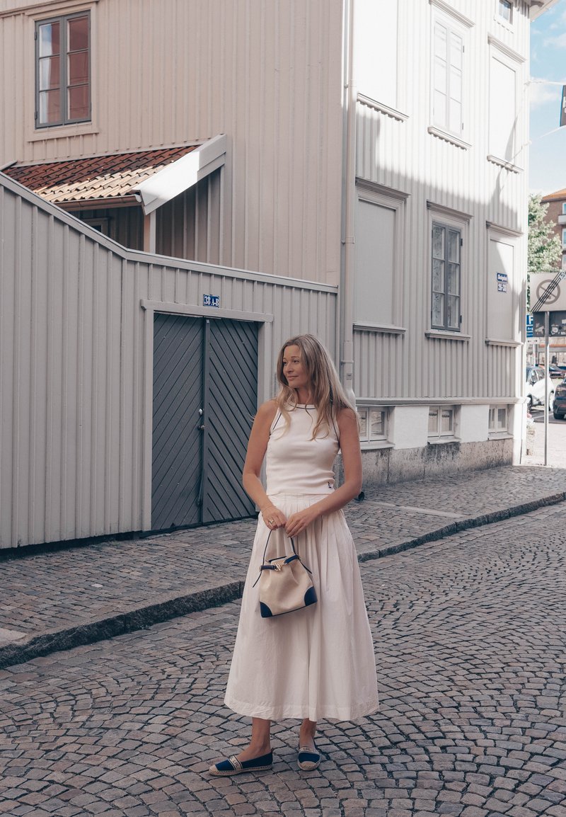 Femme en robe blanche tenant un sac à main beige, debout dans une rue pavée à côté de bâtiments en bois gris clair avec portes et fenêtres fermées.