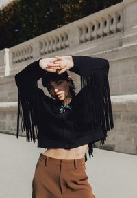Femme posant à l'extérieur, portant une veste courte noire à franges et un pantalon marron taille haute, avec la lumière du soleil projetant des ombres sur son visage et ses bras.